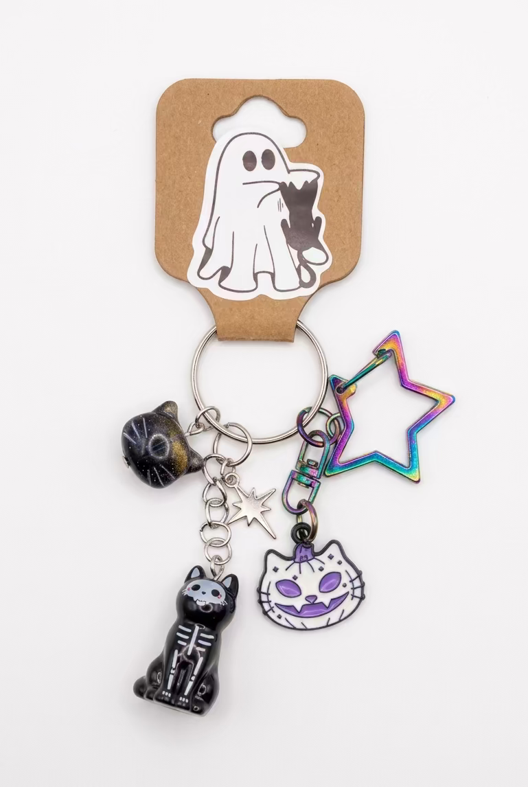 A Halloween-themed keychain with multiple charms including a 3D sitting black cat skeleton figure charm, a purple pumpkin-cat face enamel charm, an iridescent cat glass bead, a holographic star-shaped keyring clip & a small silver star charm, on silver tone keyring split ring. Photographed on a plain white surface. 
